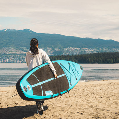 Woman walking Cascadia paddle board with blue Evo branding towards water on a Vancouver beach.