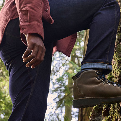 Close up torso shot of someone lifting their right foot up against the mossy bark of a tree in a forest. They are wearing hiking boots and a thick flannel shirt.